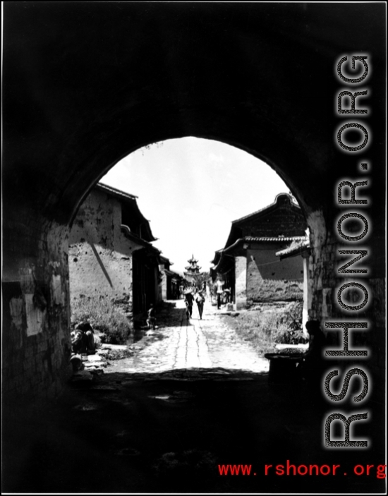 Looking into an old Chinese village through a gate, with a drum tower or bell tower in the distance, in the middle of the village. During WWII.