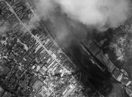 Bombing of small riverside town likely in SW China (esp. Guangxi), but possibly in Burma, or French Indochina. During WWII. Signs of previous bombings are visible about, including destroyed houses along the main road, and bomb craters here and there. Smoke arises in this image from recent bomb blasts.