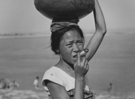 A woman carries a ceramic pot on her head, while smoking an enormous rolled cigarette. In the CBI in WWII.