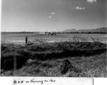 B-24 stirs up dust on runway at Kunming, China, during WWII. Chinese worker stands in the foreground, as well as some in the far distance.