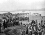 A large crowd along a river in China during WWII, gathered for some kind of communal activity. On the right is a dragon dance team, and numerous people hold banners and flags.