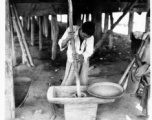 Local people in Burma near the 797th Engineer Forestry Company--A man is working most likely to dehusk grain, or to make some other worked produce, while another person waits with a woven dry pan.  During WWII.