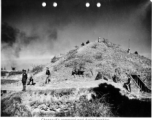 Pyramid-shaped air raid shelter used by Chennault in Kunming during WWII, in the midst of an air raid. In this image, Chennault is standing toward left, looking at the Japanese bombing out of scene of the left. In the middle, an American GI makes plots on a map board. Air vents can be seen protruding from the top, and garden vegetables grow in front.  Note also the female professional staff. A woman, probably a westerner, sits next to the GI with the map board, and another woman, likely of Chinese descent, 
